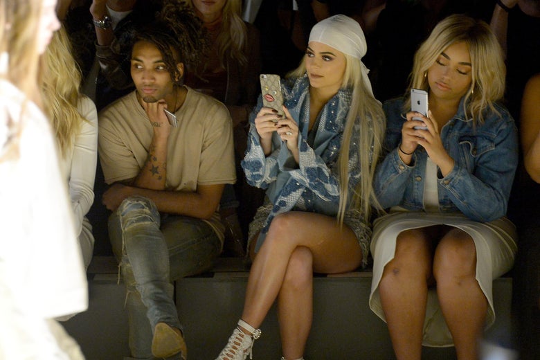 Kylie Jenner (C) and Jordyn Woods (R) sit front row at a fashion show, both taking pictures on their phone
